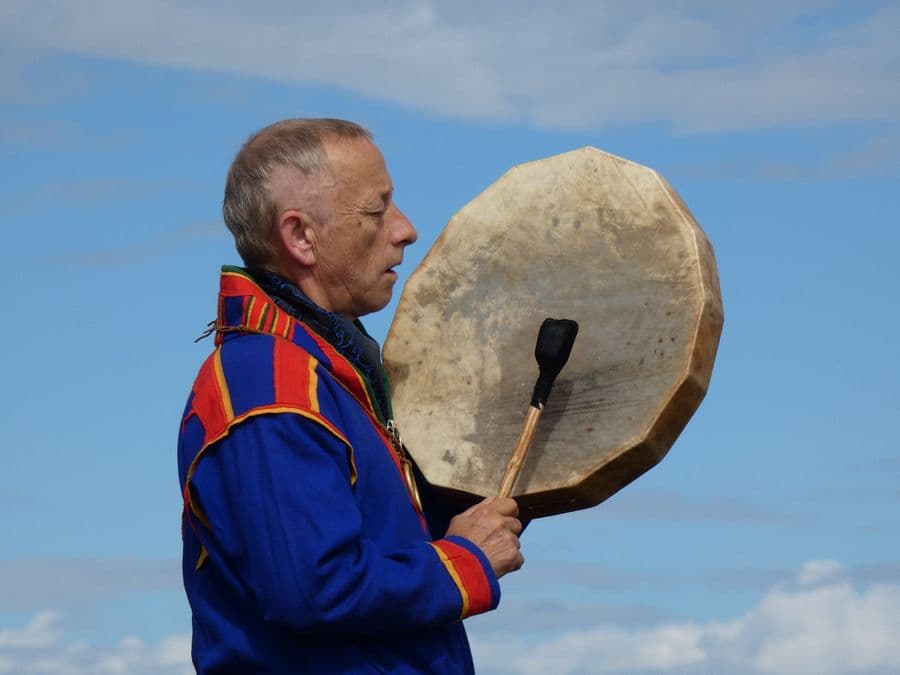 A Sámi shaman in blue gákti plays the frame drum under an open sky